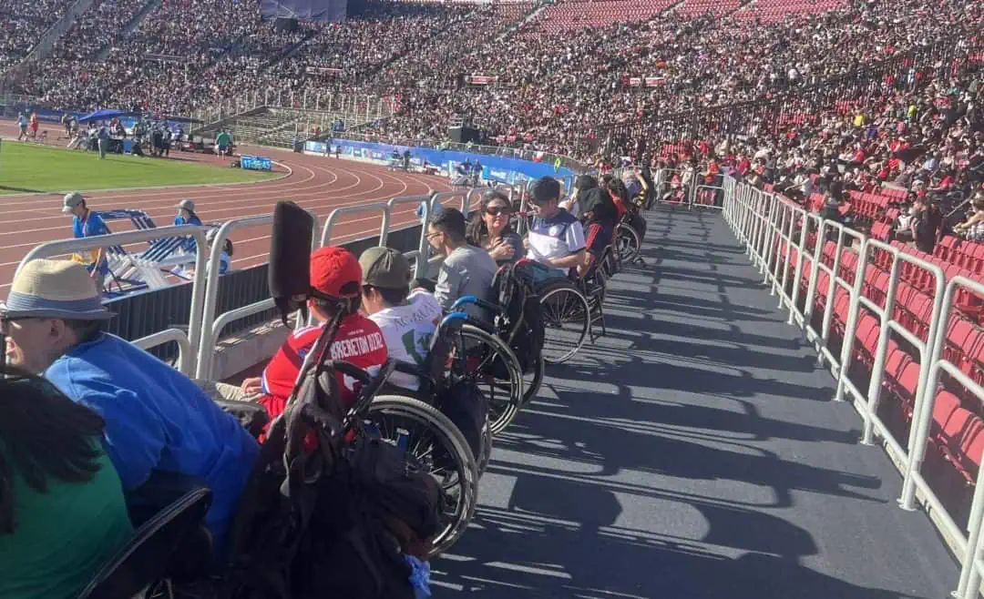 Fotografía tomada en una tribuna del estadio nacional donde aparecen varias personas en silla de ruedas mirando un partido desde una plataforma plana que hay en la parte baja de la tribuna.