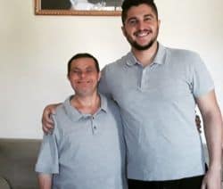 Un hombre de aproximadamente 60 años y con síndrome de down está abrazado a su hijo de unos 30 años. Ambos sonríen a la cámara.