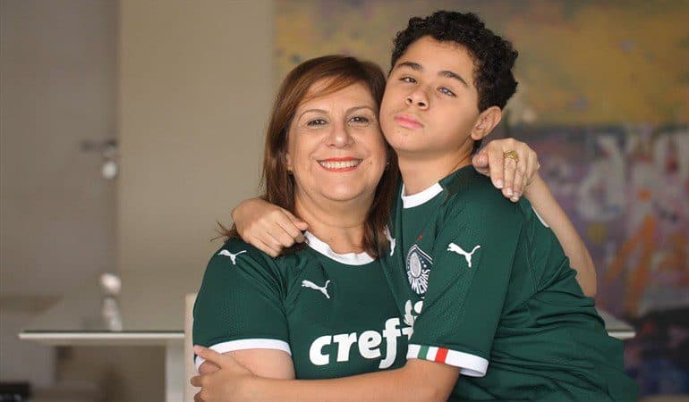 Una madre y su hijo ciego se abrazan y sonríen a la cámara en su casa. Ambos están con la camiseta verde del club de fútbol Palmeiras de Brasil.