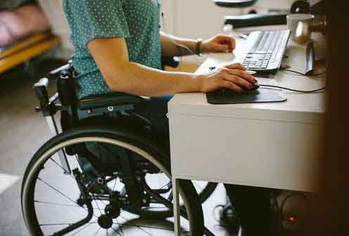 Hay una persona en silla de ruedas trabajando en su computador en su casa.