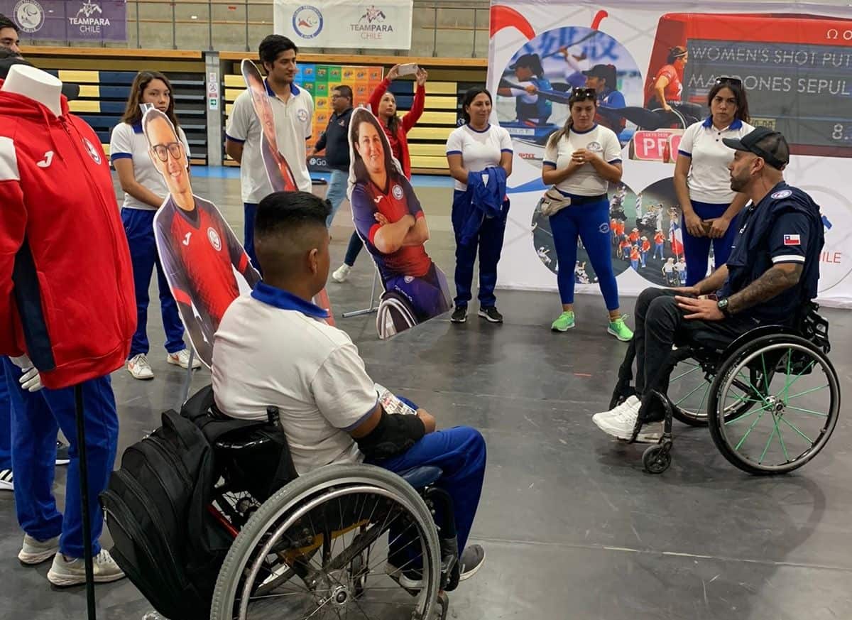 Foto de un grupo de personas en silla de ruedas dentro de una cancha techada mientras uno de ellos le habla al resto.