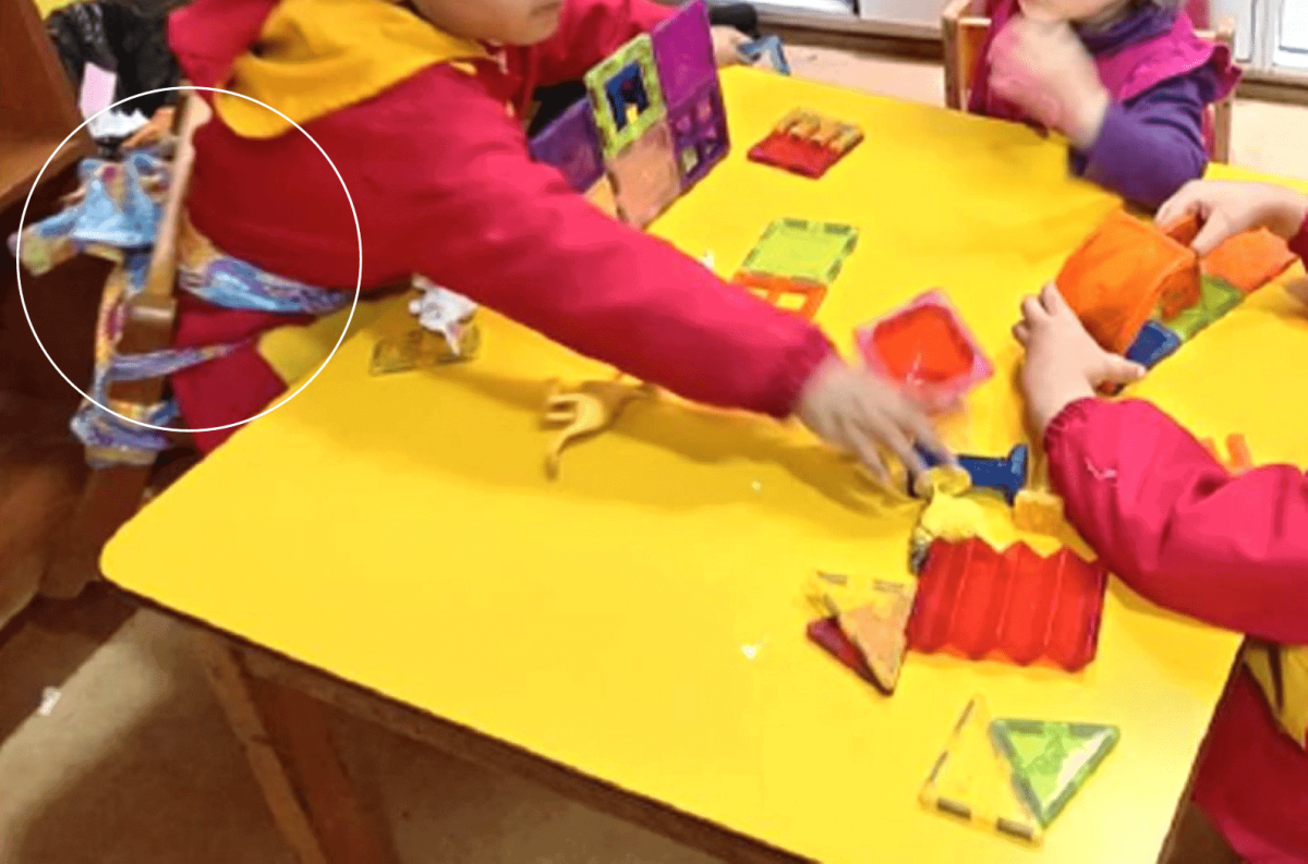 Foto dentro de una sala de una jardín infantil, donde hay tres niños sentados en una mesa, y uno de ellos está amarrado a la silla a través de unas telas que rodean su cintura.