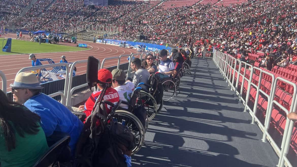 Fotografía tomada en una tribuna del estadio nacional donde aparecen varias personas en silla de ruedas mirando un partido desde una plataforma plana que hay en la parte baja de la tribuna.