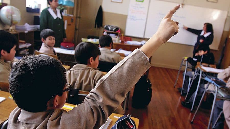 Foto dentro de una sala de clases de escuela básica, donde aparece un niño de espaldas levantando la mano.