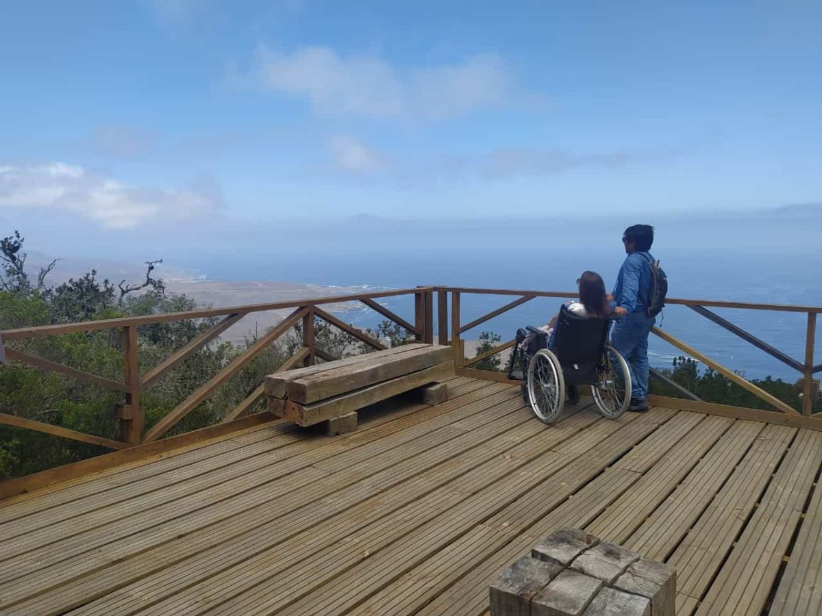 Foto donde aparece una persona en silla de ruedas y otra de pie a su lado y están en un mirador con vista hacia el mar.