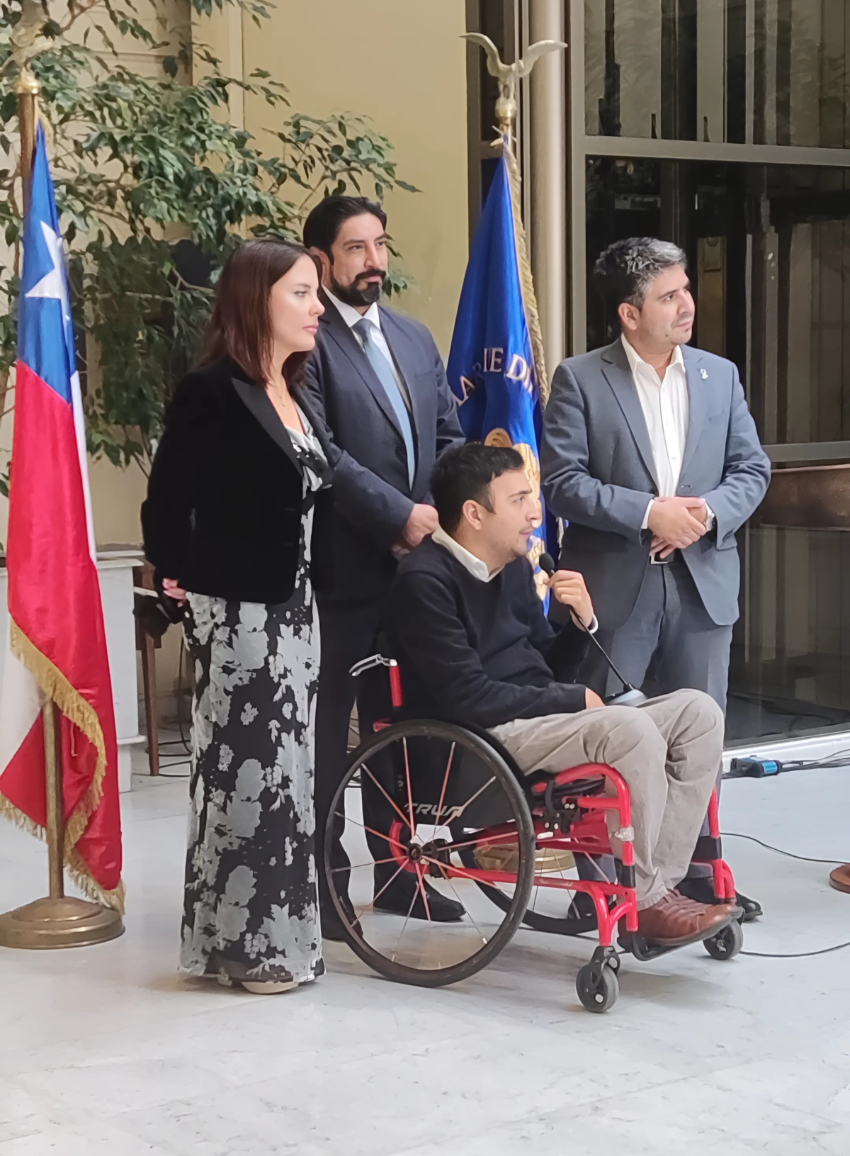 Foto en el congreso nacional donde aparecen 3 diputados de pie junto a una persona en silla de ruedas hablando a la prensa.