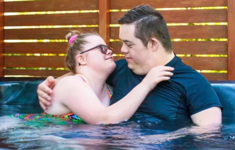 Una pareja de personas con síndrome de Down se abrazan y se miran a los ojos mientras se están bañando en un jacuzzi.