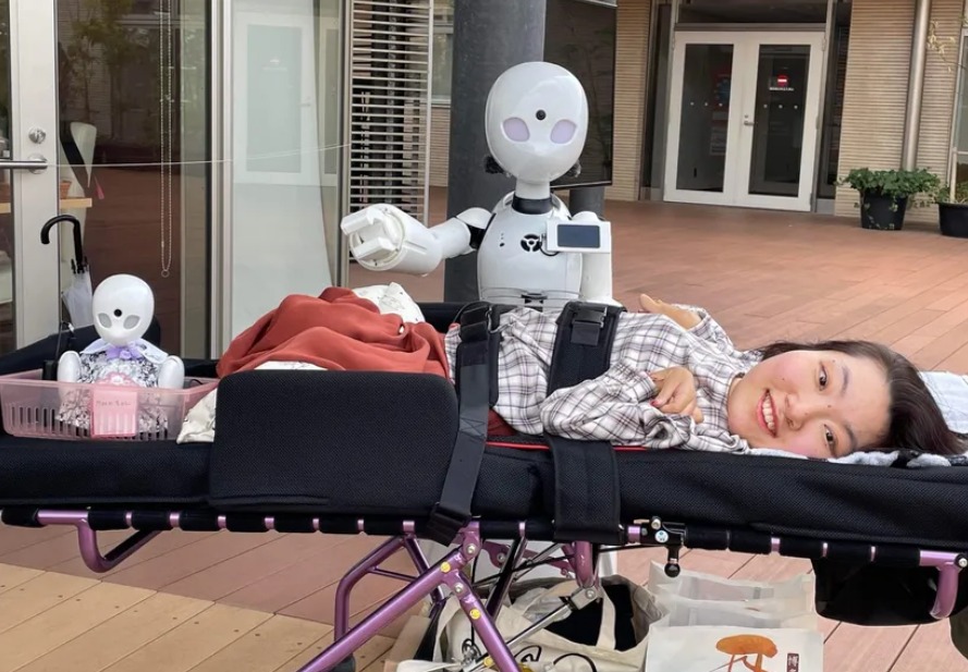 FOto de una persona con paralisis cerebral acostada en una cama, y a su lado está uno de los robots que ella puede manejar para trabajar en la cafetería.