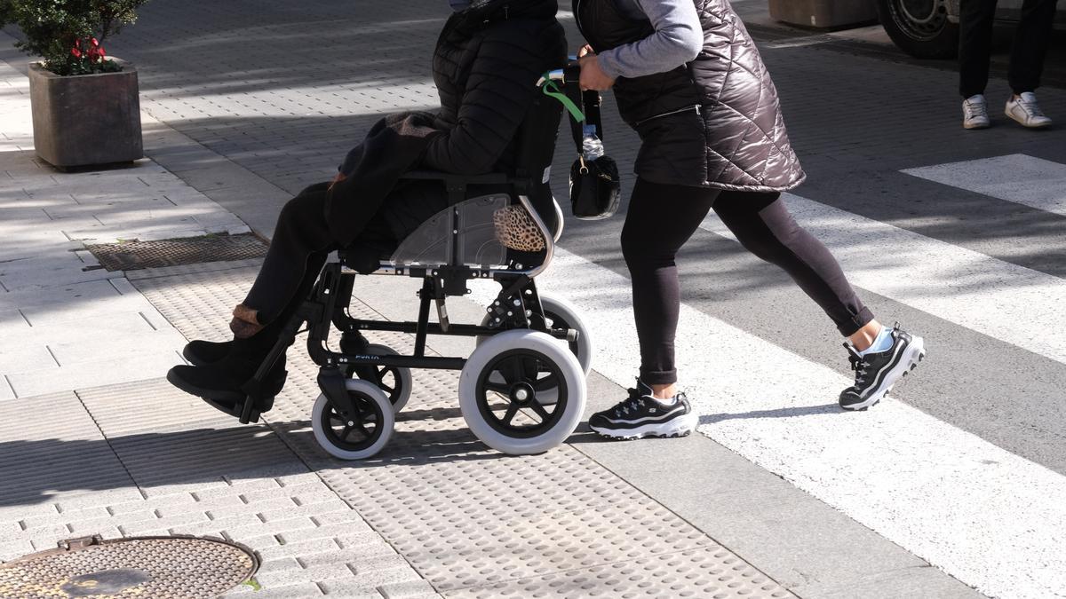 Foto de una persona llevando a otra persona en silla de ruedas por la vereda en una ciudad