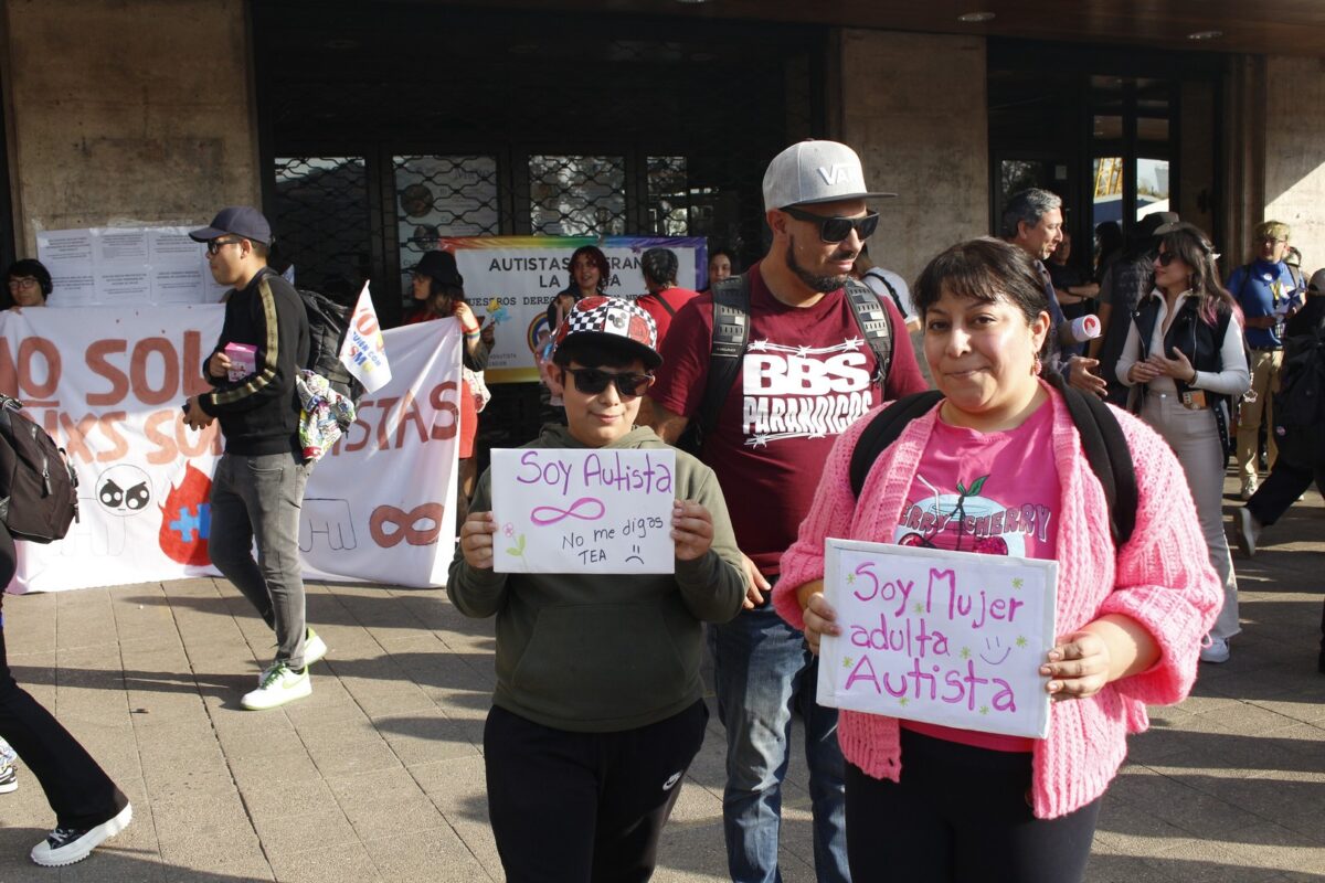 Foto de un niño junto a su madre durante una marcha de personas autistas, y el niño sostiene un cartel que dice soy autista no me digas tea