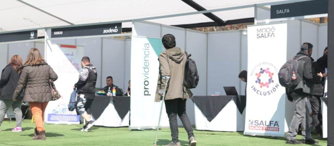 Foto de una persona con muletas caminando en una feria laboral donde hay varios puestos de diferentes empresas y organizaciones.