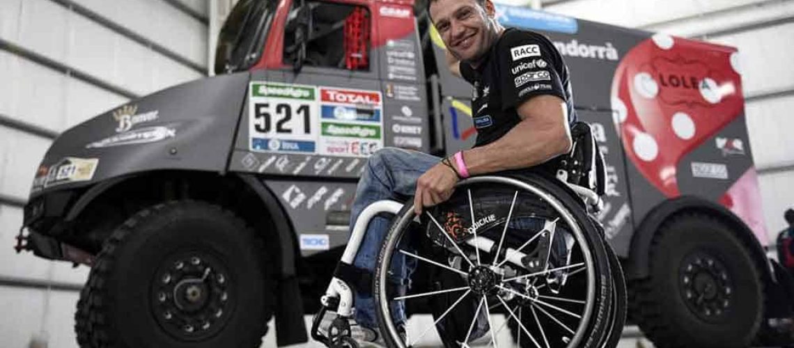 El piloto Albert Llovera sonriendo en su silla de ruedas y atrás se ve el enorme camión de carreras que conduce.