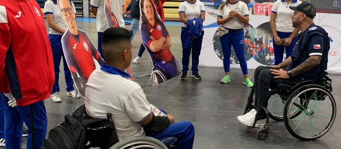 Foto de un grupo de personas en silla de ruedas dentro de una cancha techada mientras uno de ellos le habla al resto.