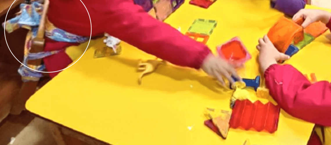 Foto dentro de una sala de una jardín infantil, donde hay tres niños sentados en una mesa, y uno de ellos está amarrado a la silla a través de unas telas que rodean su cintura.