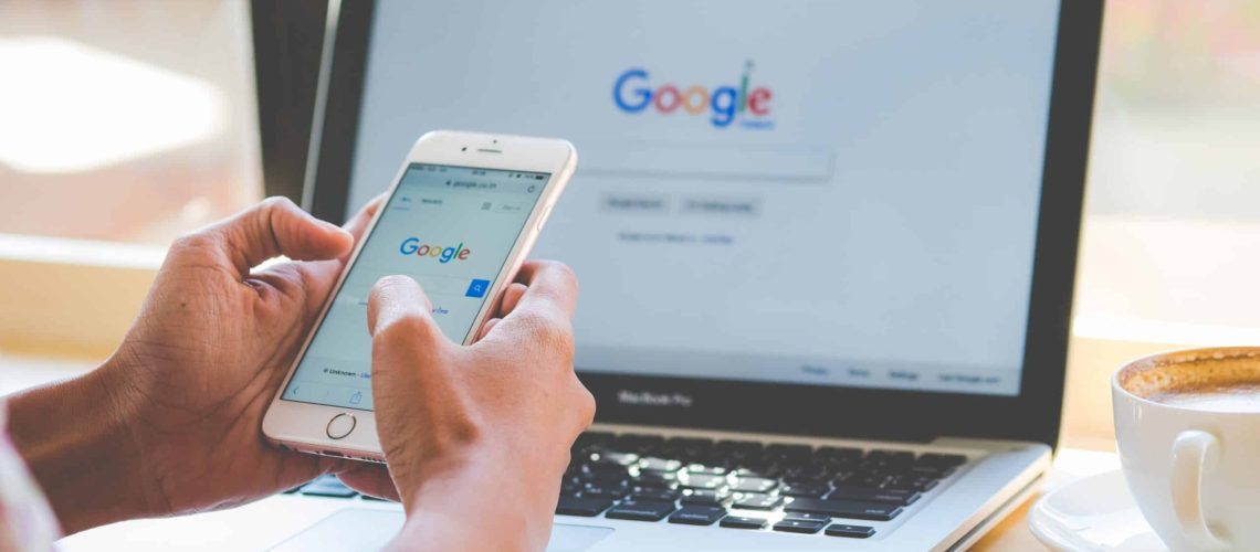 Foto donde aparecen las manos de una persona sosteniendo un teléfono y frente a ella en un escritorio hay un computador abierto, en ambas pantallas se ve la portada de google