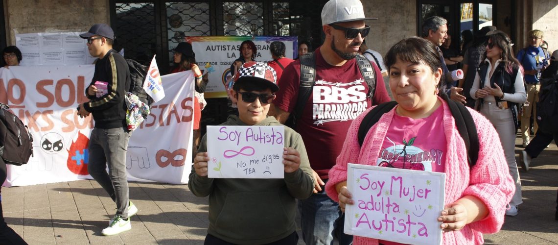 Foto de un niño junto a su madre durante una marcha de personas autistas, y el niño sostiene un cartel que dice soy autista no me digas tea