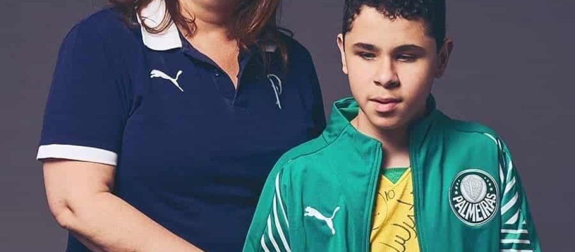 Una mujer abrazando a su hijo en un estudio de fotografía. Ella tiene una camiseta azul del club de fútbol Palmeiras de Brasil y su hijo, quien es ciego, un polerón verde del mismo club.