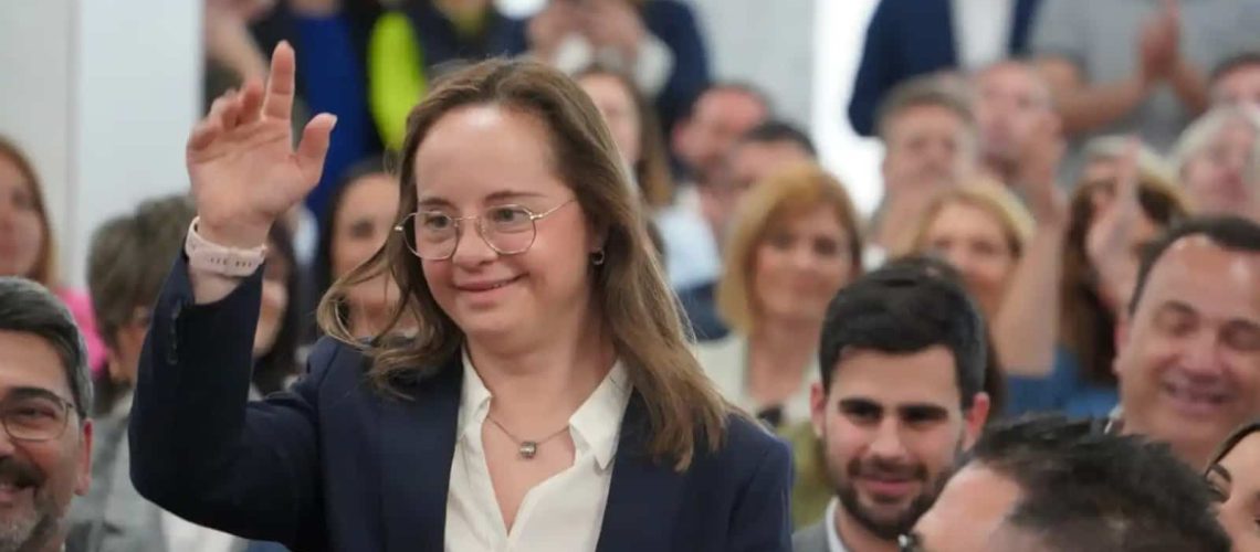 Foto en un salón donde aparece una mujer con síndrome de Down quien viste tenida formal y saluda con una mano en alto a la gente que le aplaude. Usa lentes y está sorniendo.