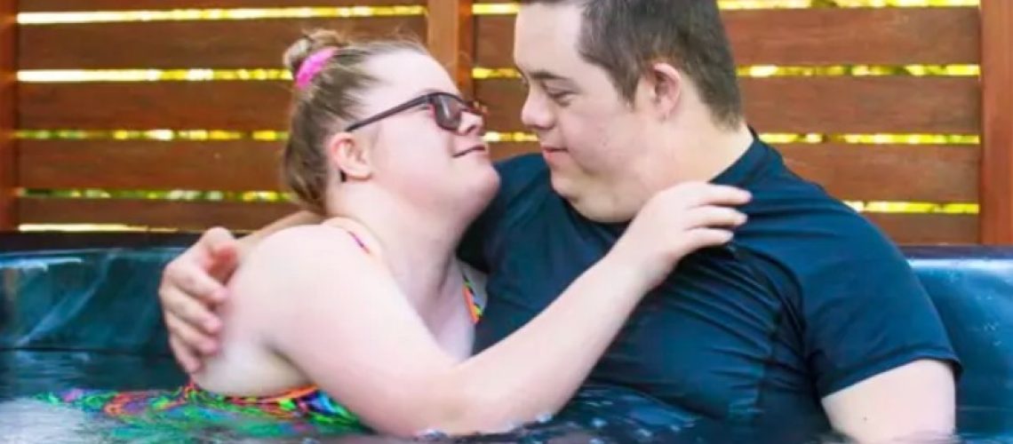 Una pareja de personas con síndrome de Down se abrazan y se miran a los ojos mientras se están bañando en un jacuzzi.