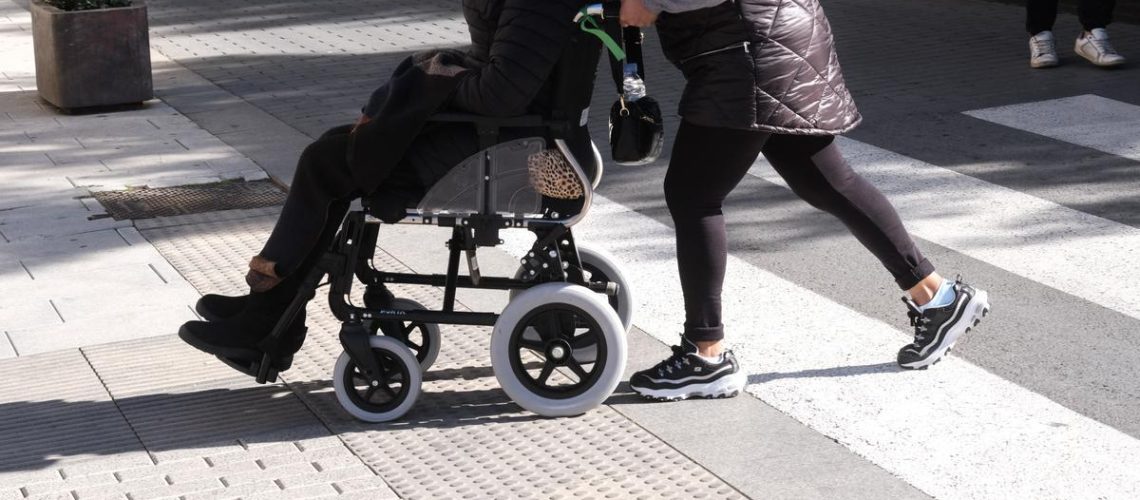 Foto de una persona llevando a otra persona en silla de ruedas por la vereda en una ciudad