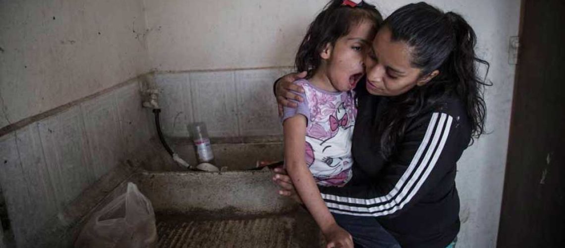 Foto de una mujer joven tomando en brazos a su hija con discapacidad dentro de una cocina que se nota muy precaria.