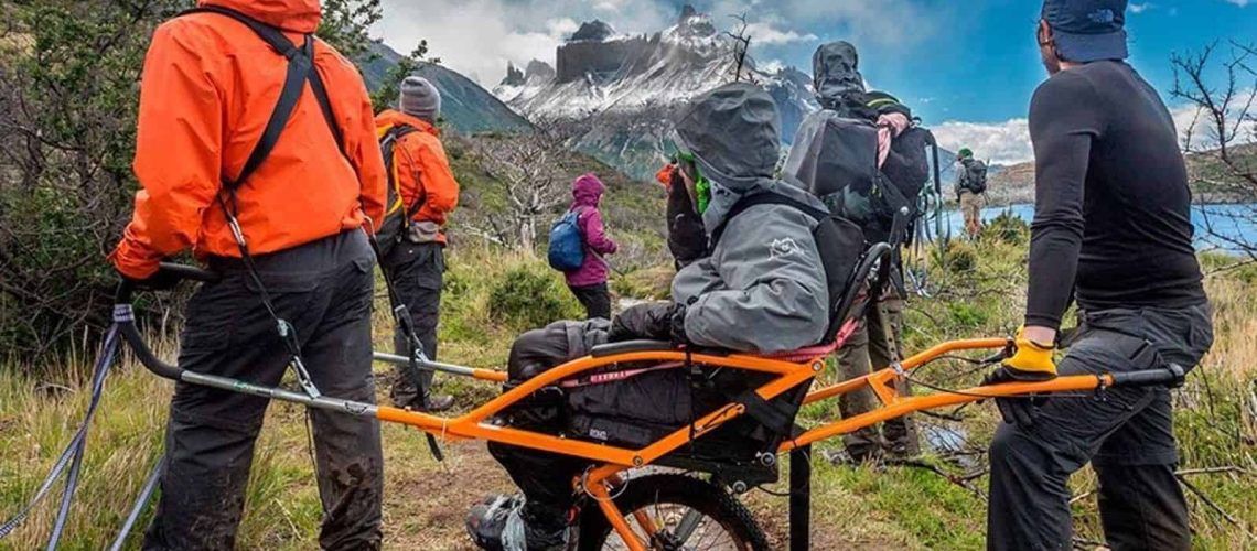 Foto en la patagonia chilena con un paisaje con harta vegetación, una laguna al lado y de fondo un cerro con nieve. Aparecen 4 personas mirando hacia el cerro, 1 de ellas está en silla de ruedas.