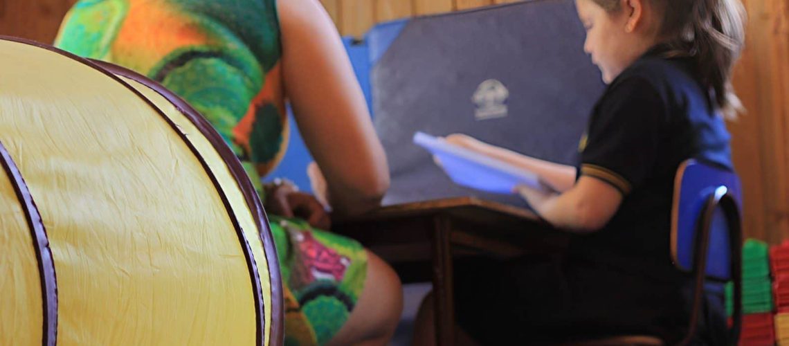 Foto en una sala de clases donde aparece una profesora junto a una alumna pequeña sentadas frente a un computador.