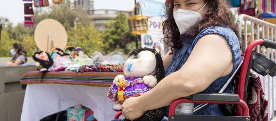 Foto en una feria artesanal donde aparece una mujer en silla de ruedas con una muñeca en la mano y atrás una mesa con más muñecas hechas por ella.