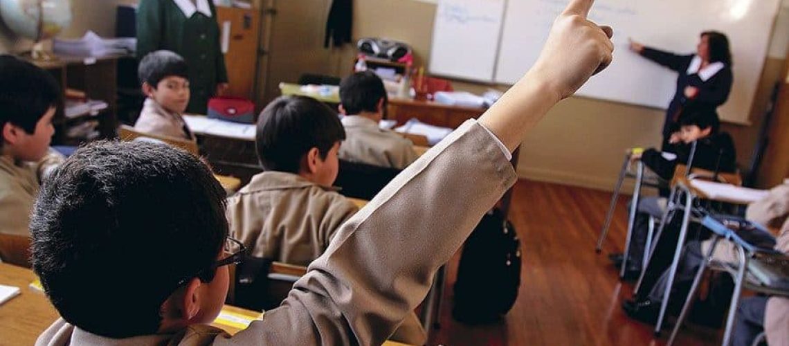 Foto dentro de una sala de clases de escuela básica, donde aparece un niño de espaldas levantando la mano.
