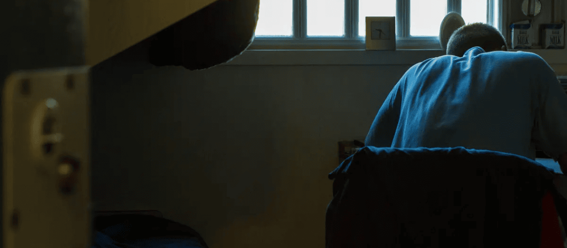 Foto dentro de una celda donde aparece una persona sentada en una silla y escribiendo algo en un escritorio. De fondo se ve una ventana con rejas por donde entra la luz a la celda.