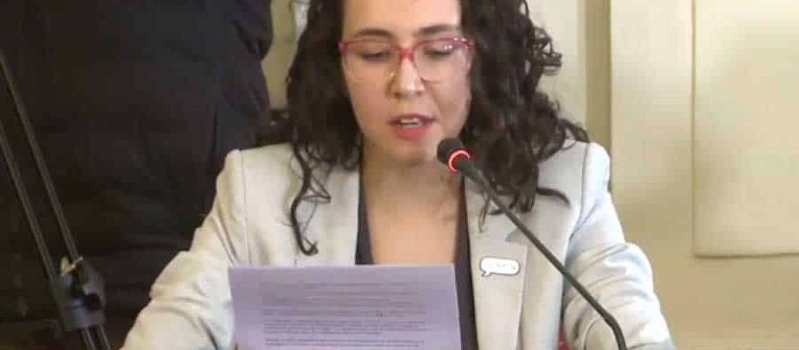 foto donde aparece una mujer autista vestida con chaqueta y sentada en una sala del senado hablando a través del micrófono mientras lee su discurso.