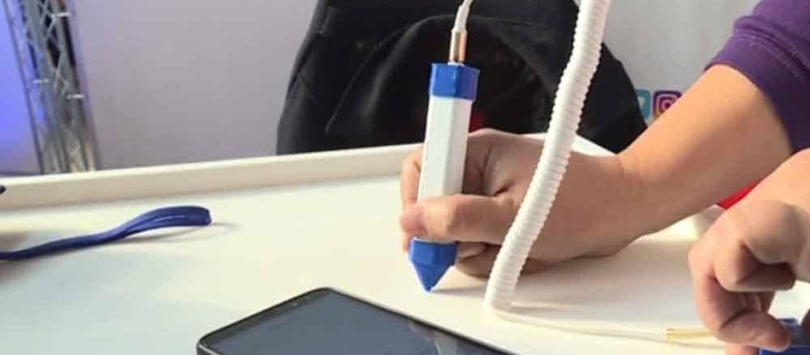 Aparece la mano de una persona escribiendo sobre una tabla con un lápiz especial, y al lado hay un celular donde se va viendo en la pantalla la traducción.