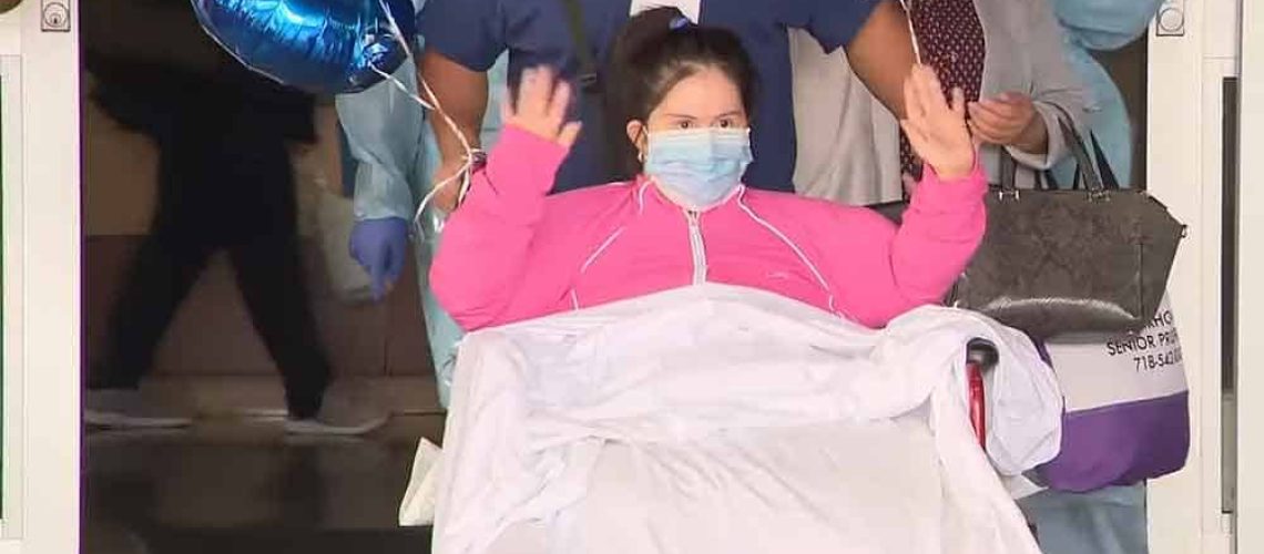 Foto de una mujer con síndrome de Down saliendo de un hospital en silla de ruedas y saludando a quienes la aplauden.