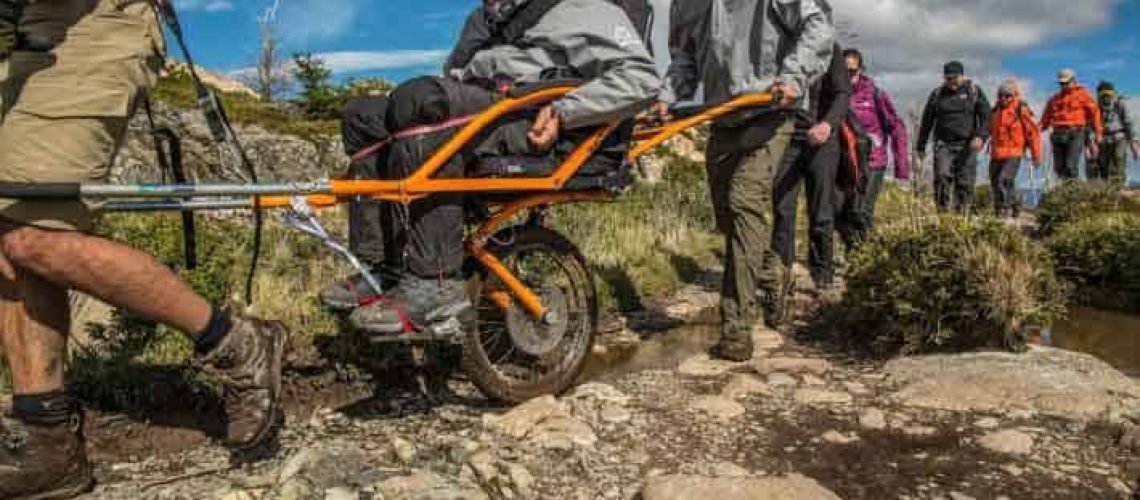 Un joven en silla de ruedas recorriendo un sendero en Torres del Paine, mientras otras personas los ayudan levantando su silla.