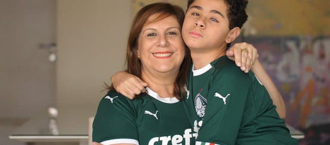 Una madre y su hijo ciego se abrazan y sonríen a la cámara en su casa. Ambos están con la camiseta verde del club de fútbol Palmeiras de Brasil.