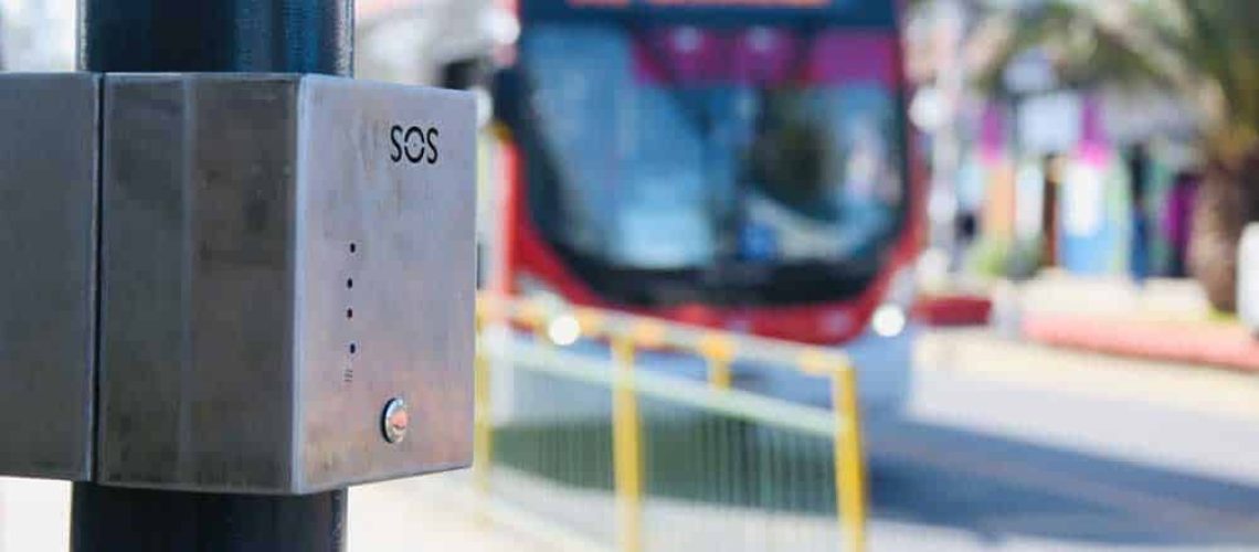 Foto de un paradero del transantiago, y en primer plano hay una caja de metal que es un botón de pánico que cuenta con sistema braille. De fondo se ve una micro llegando al paradero.