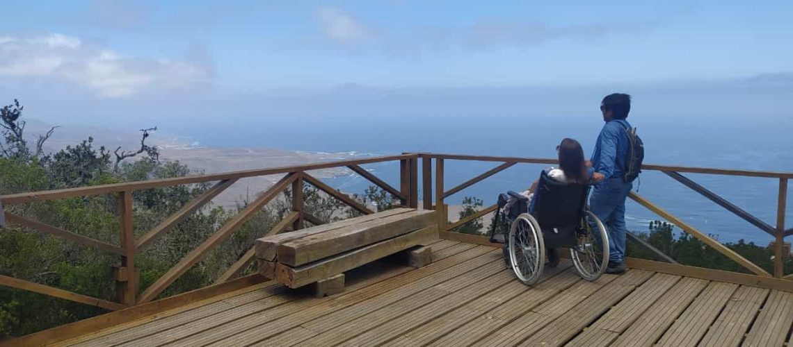 Foto donde aparece una persona en silla de ruedas y otra de pie a su lado y están en un mirador con vista hacia el mar.