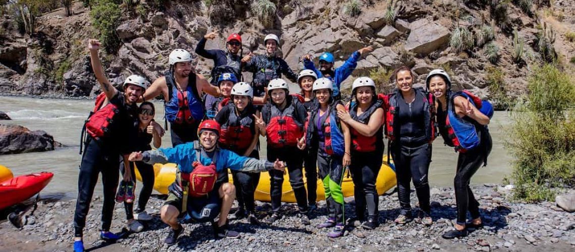 Foto de un grupo de personas sordas junto a interprete de lengua de señas y guía turístico, al borde de un rio, con el cerro de fondo, y con trajes de agua sonriendo a la cámara.