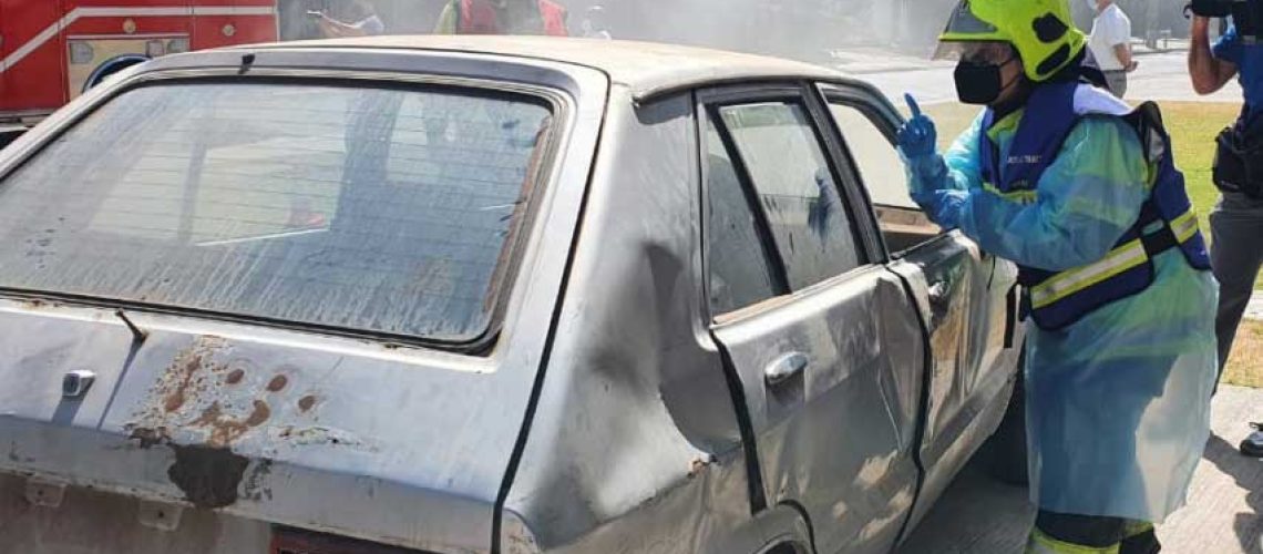 Foto de un simulacro de bomberos donde aparece una bombera comunicandose en señas con una persona dentro de un auto accidentado.