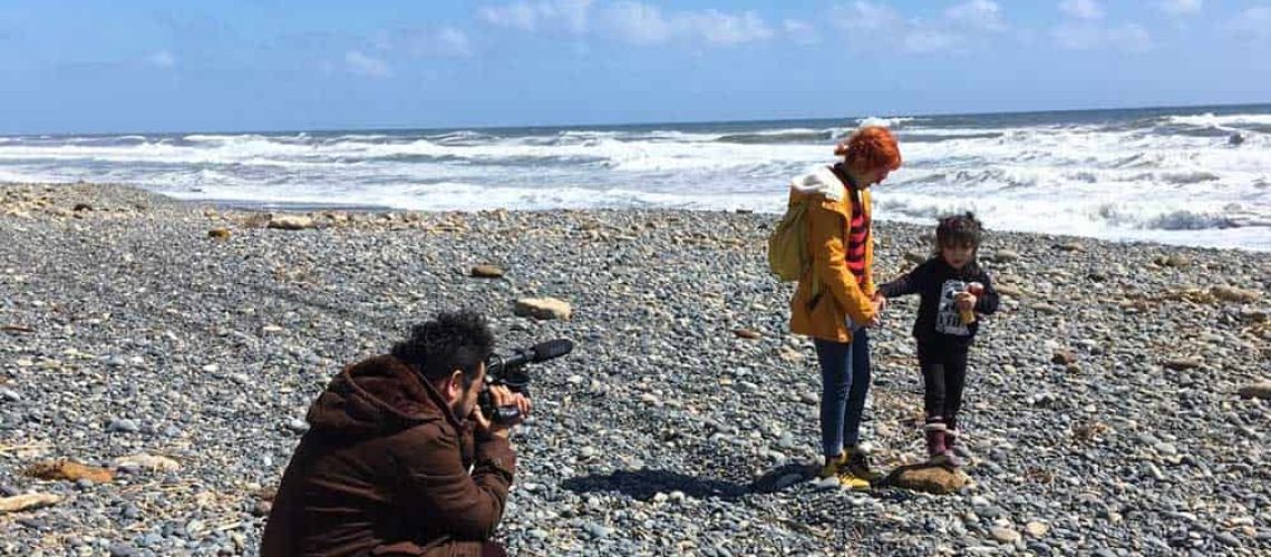 Foto en una playa grande y vacía en un día soleado en el sur de Chile, donde aparece una mujer de su mano con su hija caminando por la orilla de la playa, y al lado una persona que va grabando todo.