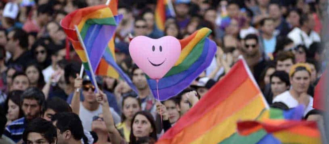 una marcha con muchas personas con banderas LGTBIQ+ (bandera de arcoiris). También aparece un globo rosado con forma de corazón y tiene dibujada una cara sonriente.