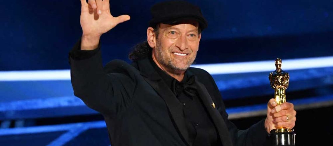 Foto del actor Troy Kotsur quien está sobre el escenario con el premio en una mano y con la otra mano hace la seña de Te Amo.