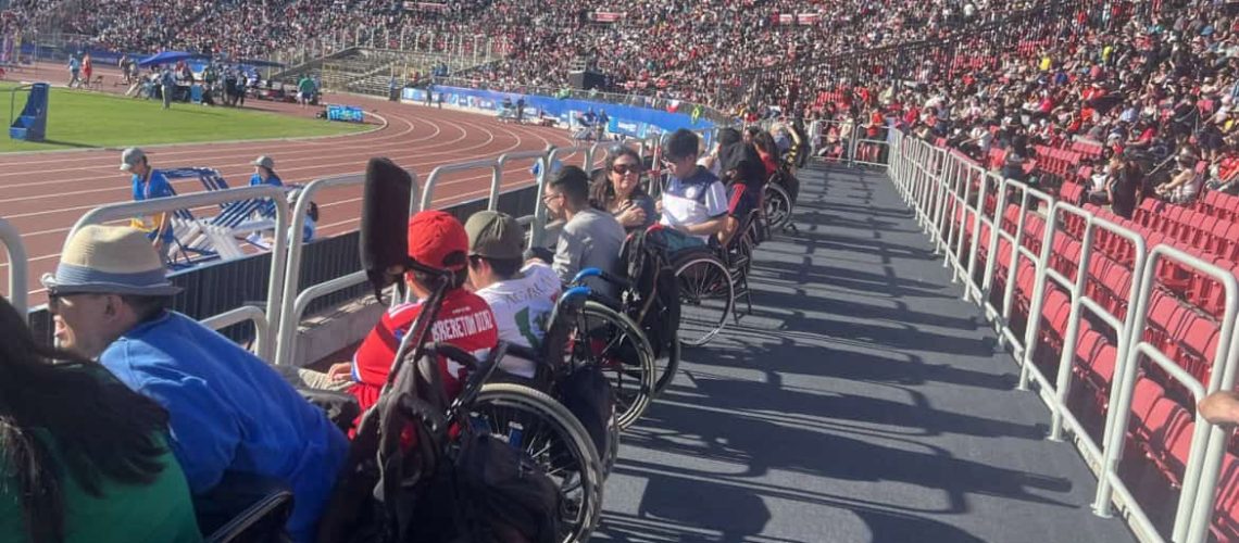 Fotografía tomada en una tribuna del estadio nacional donde aparecen varias personas en silla de ruedas mirando un partido desde una plataforma plana que hay en la parte baja de la tribuna.