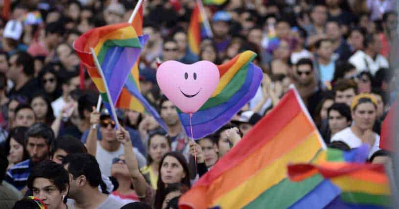 una marcha con muchas personas con banderas LGTBIQ+ (bandera de arcoiris). También aparece un globo rosado con forma de corazón y tiene dibujada una cara sonriente.