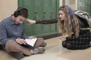 El protagonista de la serie aparece sentado en un pasillo de la escuela, mirando un cuaderno, con audífonos puestos y una compañera tocándole el hombro para que la mire.